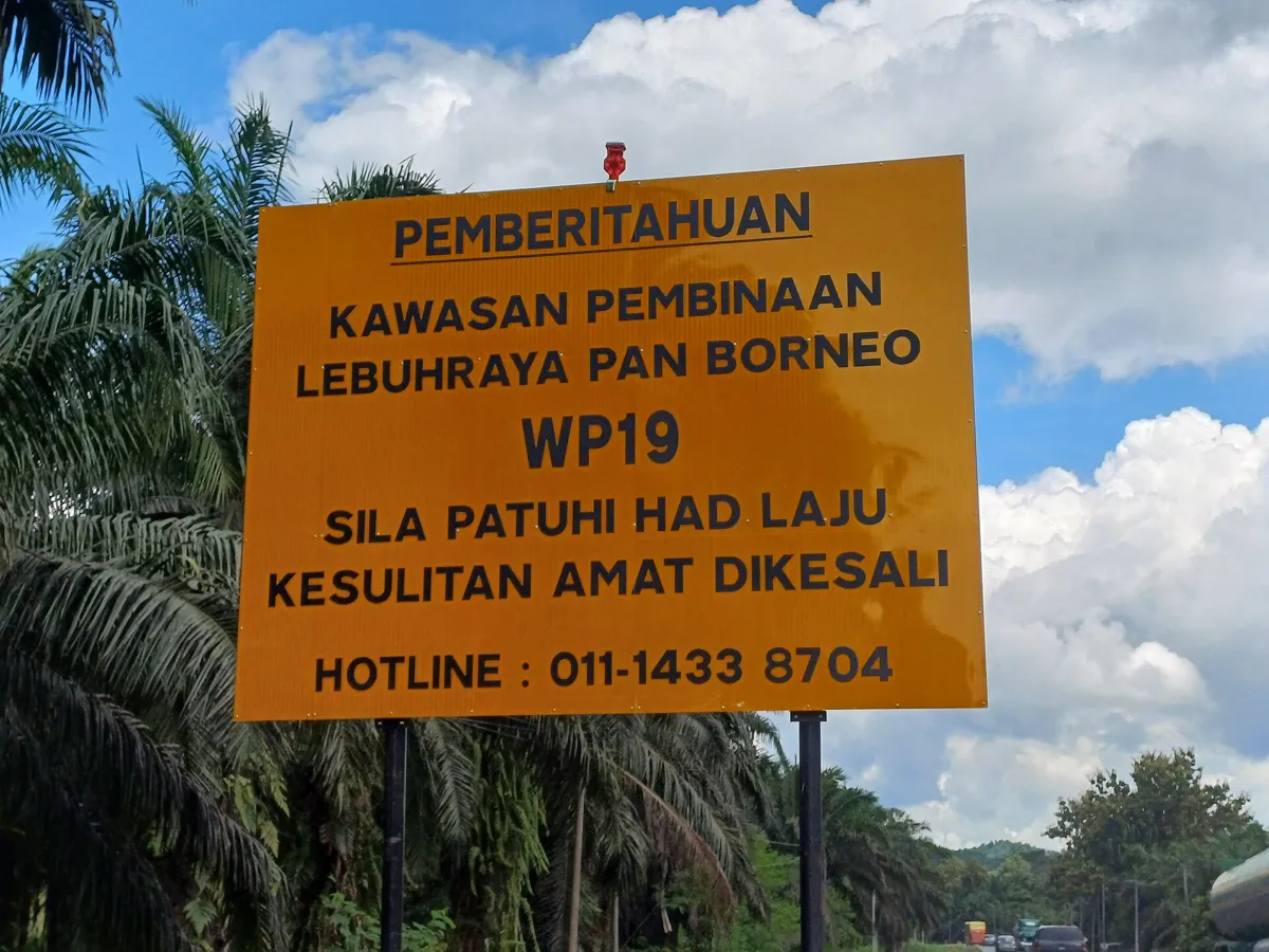 Yellow roadside notice reading Kawasan Pembinaan Lebuhraya Pan Borneo WP19, with oil palms behind